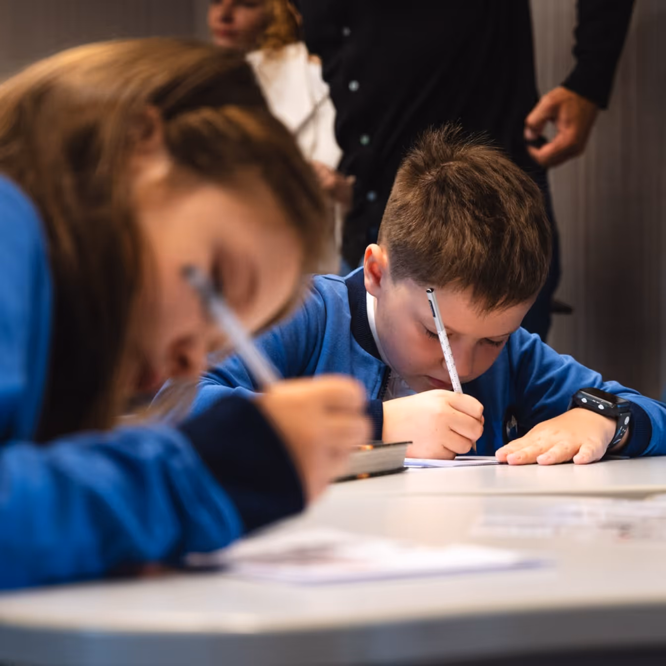 Two Erudito Lyceum students in blue uniforms writing at a table, adults blurred in the background.