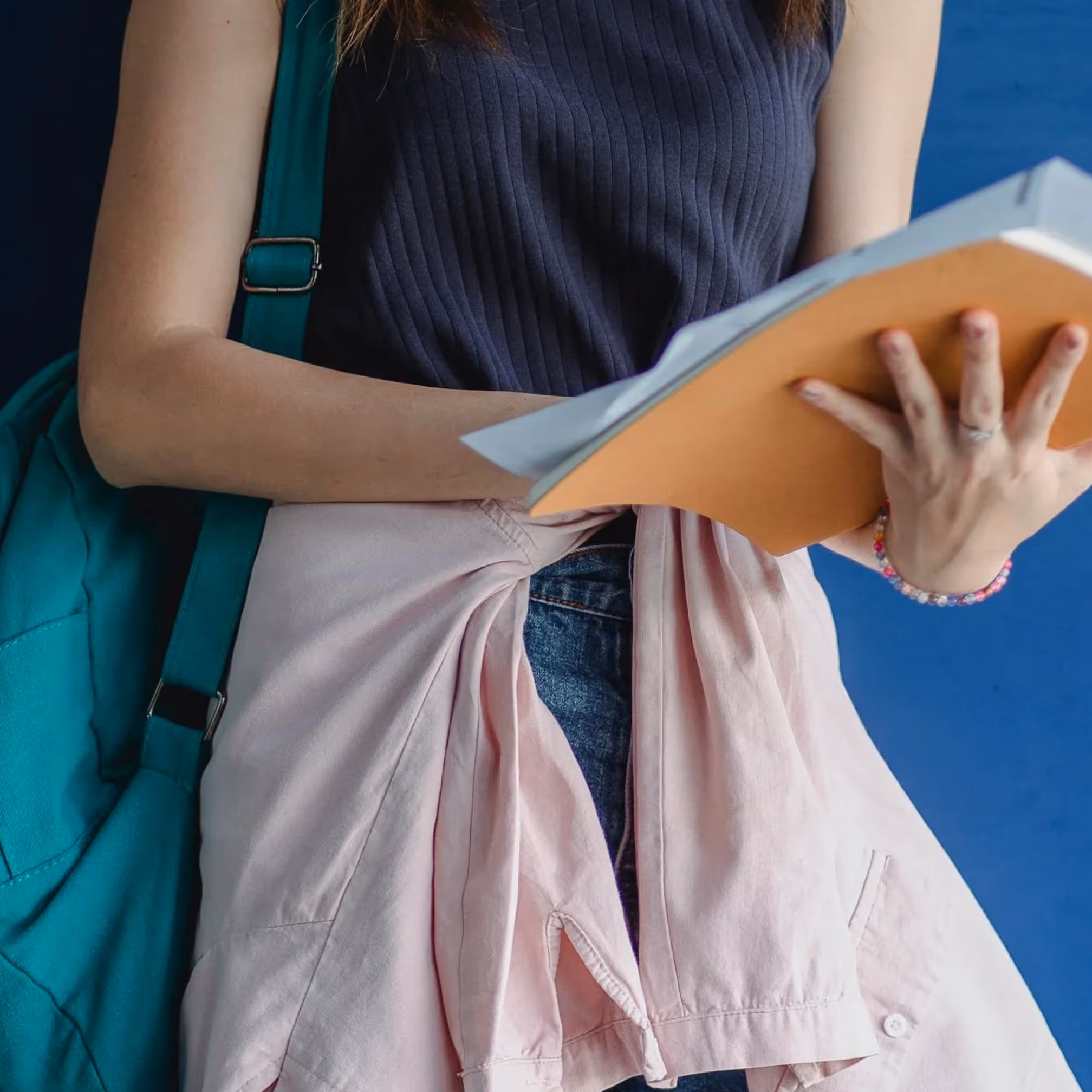 Close-up of a student holding a notebook and folder, teal backpack strap over her shoulder, against blue.